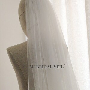 Pu&ograve; includere: Un velo da sposa bianco e trasparente &egrave; drappeggiato sulla testa e sulle spalle di un manichino. Il velo &egrave; lungo e fluente, con una trama delicata. Il testo "MI BRIDAL VEIL" &egrave; stampato in nero sulla spalla del manichino.
