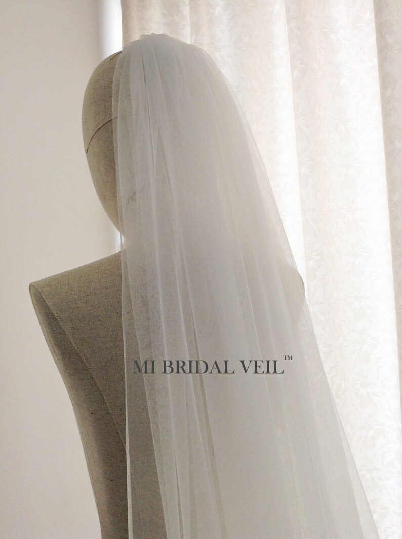 Pu&ograve; includere: Un velo da sposa bianco e trasparente &egrave; drappeggiato sulla testa e sulle spalle di un manichino. Il velo &egrave; lungo e fluente, con una trama delicata. Il testo "MI BRIDAL VEIL" &egrave; stampato in nero sulla spalla del manichino.