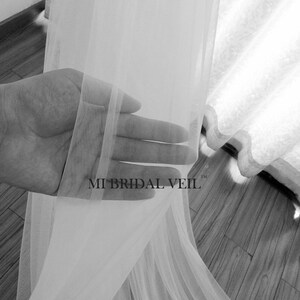 Pu&ograve; includere: Un velo da sposa bianco trasparente &egrave; tenuto in mano, mostrando il suo tessuto delicato e fluido. Il velo &egrave; semitrasparente, lasciando passare la luce. Il testo "MI BRIDAL VEIL" &egrave; visibile sul velo.