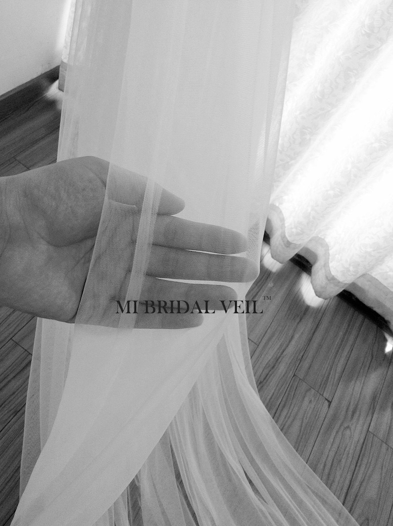 Pu&ograve; includere: Un velo da sposa bianco trasparente &egrave; tenuto in mano, mostrando il suo tessuto delicato e fluido. Il velo &egrave; semitrasparente, lasciando passare la luce. Il testo "MI BRIDAL VEIL" &egrave; visibile sul velo.