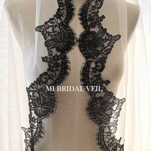 May include: A bridal veil with intricate black lace detailing. The lace forms a decorative pattern along the edges and features floral designs. The veil is sheer, allowing light to pass through, and is displayed on a mannequin.