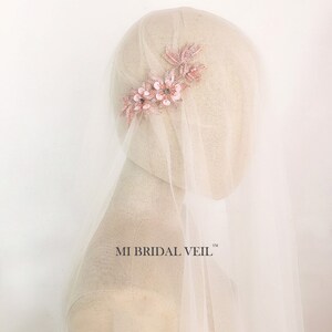 May include: A bridal veil with a delicate floral hairpiece. The hairpiece features pink fabric flowers and leaves with sparkling accents. The veil is a sheer, off-white fabric. The text "MI BRIDAL VEIL" is visible.