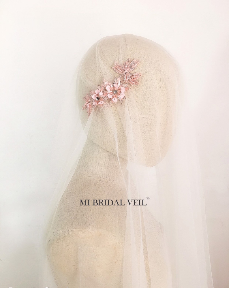 May include: A bridal veil with a delicate floral hairpiece. The hairpiece features pink fabric flowers and leaves with sparkling accents. The veil is a sheer, off-white fabric. The text "MI BRIDAL VEIL" is visible.