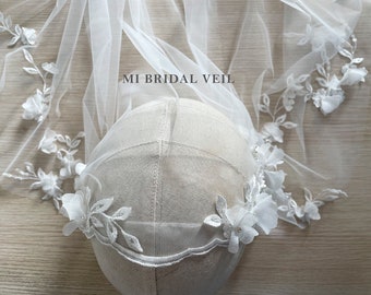 Puede incluir: Un velo de novia blanco con adornos florales. El velo, hecho de tela transparente, presenta delicadas flores y hojas tridimensionales en los bordes. El texto "MI BRIDAL VEIL" es visible en el velo.