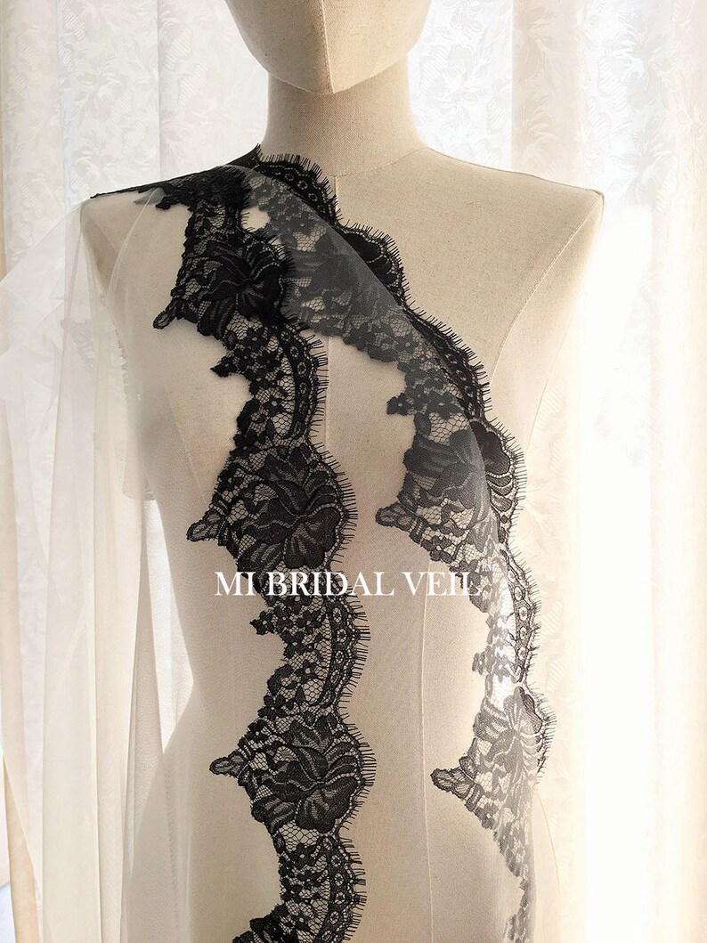 May include: A bridal veil with a delicate black lace trim. The lace features intricate floral patterns and a scalloped edge. The veil is displayed on a mannequin, showcasing its design and texture. The text "MI BRIDAL VEIL" is visible.