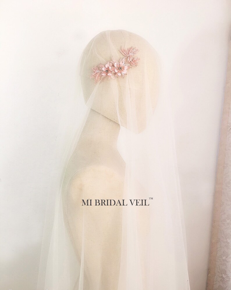 May include: A bridal veil with a delicate floral hair comb. The comb features pink fabric flowers and leaves with small embellishments. The veil is long and sheer, draping over a mannequin. The text "MI BRIDAL VEIL" is visible.