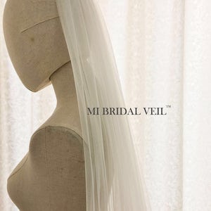 May include: A sheer, white bridal veil is draped over a mannequin. The veil is long and flowing, with a delicate texture. The text "MI BRIDAL VEIL" is printed in black on the right side of the image. The background is a white, patterned fabric.