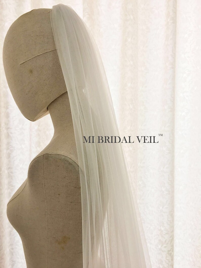 May include: A sheer, white bridal veil is draped over a mannequin. The veil is long and flowing, with a delicate texture. The text "MI BRIDAL VEIL" is printed in black on the right side of the image. The background is a white, patterned fabric.
