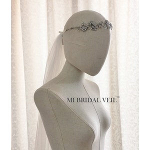 Può includere: Un copricapo da sposa argentato con un design floreale e cristalli, abbinato a un velo bianco trasparente. Il velo è attaccato con un nastro. Il testo "MI BRIDAL VEIL" è visibile. Presentato su un manichino.