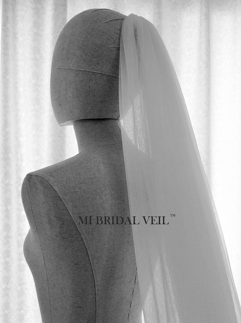 Pu&ograve; includere: Un velo da sposa bianco trasparente &egrave; drappeggiato su un manichino. Il velo &egrave; lungo e fluente, con una consistenza morbida e delicata. Il manichino &egrave; grigio e ha uno sfondo neutro. Il testo "MI BRIDAL VEIL" &egrave; visibile.