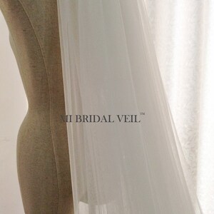 Pu&ograve; includere: Un velo da sposa bianco trasparente drappeggiato su un manichino. Il velo &egrave; realizzato in un tessuto leggero e fluido. Il testo "MI BRIDAL VEIL" &egrave; visibile sul velo. Lo sfondo &egrave; una tenda bianca.