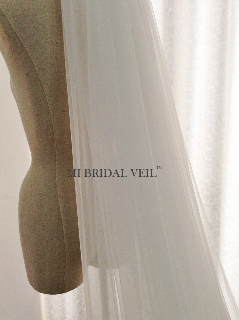 Pu&ograve; includere: Un velo da sposa bianco trasparente drappeggiato su un manichino. Il velo &egrave; realizzato in un tessuto leggero e fluido. Il testo "MI BRIDAL VEIL" &egrave; visibile sul velo. Lo sfondo &egrave; una tenda bianca.