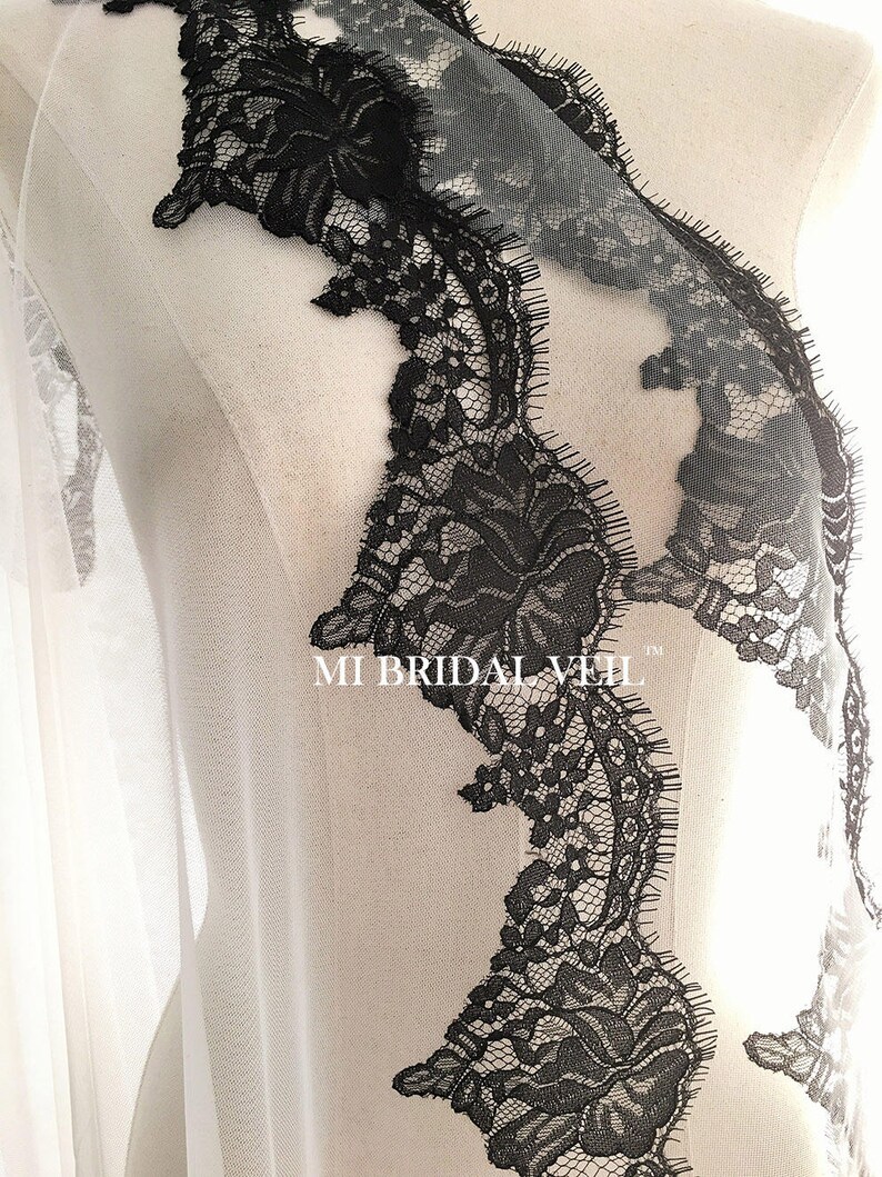 May include: Close-up of a bridal veil with intricate black lace detailing. The lace features floral patterns and a scalloped edge, creating a delicate and elegant design. The text "MI BRIDAL VEIL" is visible.