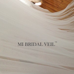 Pu&ograve; includere: Un velo da sposa bianco e trasparente che scorre. Il velo &egrave; drappeggiato, con il testo "MI BRIDAL VEIL" in basso al centro. Il tessuto appare morbido e delicato, con pieghe e trame sottili.
