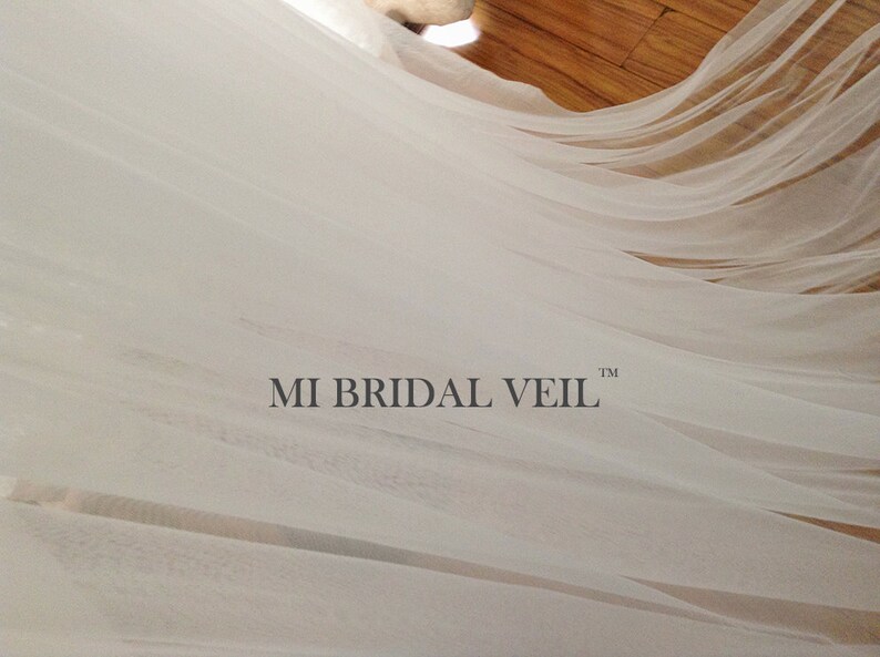 Pu&ograve; includere: Un velo da sposa bianco e trasparente che scorre. Il velo &egrave; drappeggiato, con il testo "MI BRIDAL VEIL" in basso al centro. Il tessuto appare morbido e delicato, con pieghe e trame sottili.