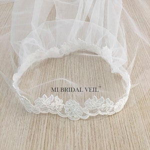 May include: A white bridal veil with a delicate floral lace trim. The veil is made of a sheer, lightweight fabric and features the text "MI BRIDAL VEIL". The lace trim is along the edge of the veil.