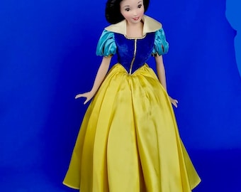 May include: A doll dressed in a yellow satin gown with a blue bodice and puffed sleeves. The doll has black hair, a red headband, and a white collar. The doll is posed against a blue backdrop.