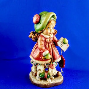 Puede incluir: Una figurita de una niña que lleva un sombrero verde con un pompón rojo, un vestido rojo y blanco, y sostiene una carta que dice "Charlie's Wish". Está de pie con dos perros pequeños.