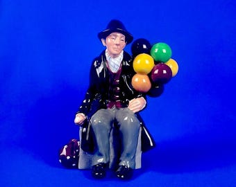 May include: A porcelain figurine of a man wearing a black coat and hat, sitting on a base and holding a bunch of colorful balloons.