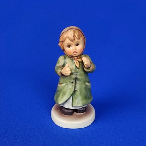 Könnte beinhalten: Eine Porzellanfigur eines jungen Mädchens, das einen grünen Mantel, einen braunen Hut und einen roten Schal trägt. Sie steht auf einem weißen Sockel.
