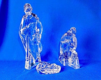 May include: A clear glass nativity set featuring three figures: a standing person, a kneeling person, and a cradle. The figures are detailed with flowing robes and are set against a blue background.