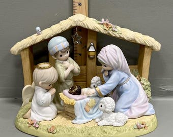 Może przedstawiać: Zestaw figurek szopki bożonarodzeniowej z porcelany, zawierający konstrukcję stajenki oraz drewnianą linijkę o wysokości około 20 cm. Zestaw zawiera figurki klęczącego anioła, stojącej postaci, matki, niemowlęcia w żłobie oraz dwóch owieczek.
