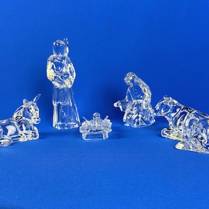 Op de afbeelding: Een heldere glazen kerststal met vijf figuren: een ezel, een staande figuur, een baby in een kribbe, een knielende figuur en een beer. De figuren staan op een blauwe achtergrond.