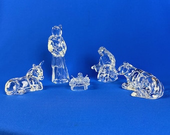 May include: A clear glass nativity set with five figures: a donkey, a standing figure, a baby in a manger, a kneeling figure, and a bear. The figures are on a blue background.