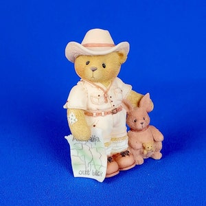 Könnte beinhalten: Figur einer Teddybär-Figur in Safari-Kleidung, mit Hut und einer Karte mit der Aufschrift "Australia out back". Eine kleine Känguru-Figur steht daneben. Die Figuren sind beige und cremefarben, vor blauem Hintergrund.