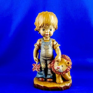 May include: A figurine of a young boy with blonde hair wearing overalls and holding a pinwheel. He is standing next to a small lion figurine.