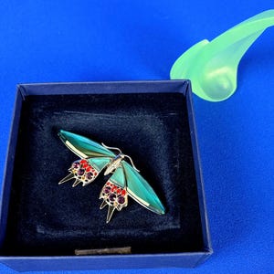 Puede incluir: Broche decorativo de mariposa con alas turquesas y detalles de gemas rojas y moradas. La broche est&aacute; colocada en una caja azul oscuro, con un objeto decorativo verde claro en el fondo.