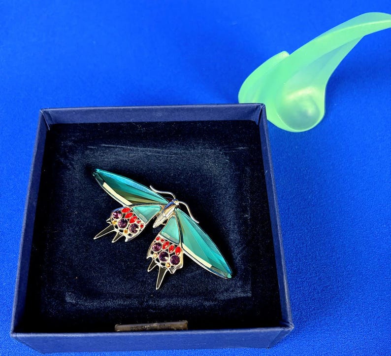 Puede incluir: Broche decorativo de mariposa con alas turquesas y detalles de gemas rojas y moradas. La broche est&aacute; colocada en una caja azul oscuro, con un objeto decorativo verde claro en el fondo.
