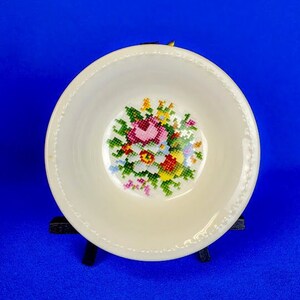 May include: A white ceramic bowl with a floral design. The bowl has a white background with a colorful embroidered floral design in the center. The bowl is on a black stand.
