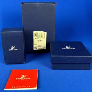 Puede incluir: Varias cajas Swarovski azul oscuro de diferentes tama&ntilde;os, con el logotipo de Swarovski en blanco. Tambi&eacute;n hay un certificado rojo con el logotipo de Swarovski. Las cajas est&aacute;n sobre un fondo azul.