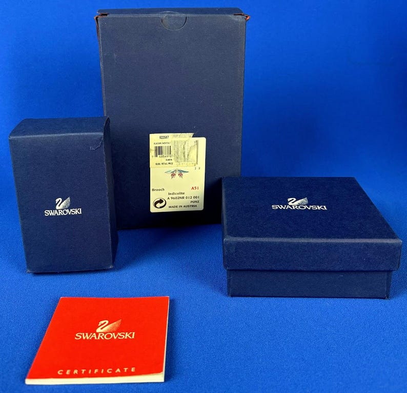 Puede incluir: Varias cajas Swarovski azul oscuro de diferentes tama&ntilde;os, con el logotipo de Swarovski en blanco. Tambi&eacute;n hay un certificado rojo con el logotipo de Swarovski. Las cajas est&aacute;n sobre un fondo azul.