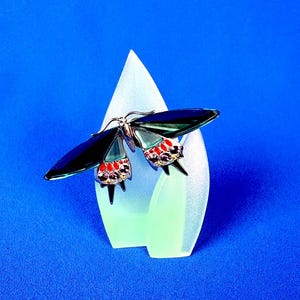 Puede incluir: Broche decorativo de mariposa con alas azul oscuro y detalles rojos y morados. El broche est&aacute; colocado sobre una pantalla en forma de l&aacute;grima verde claro sobre un fondo azul.