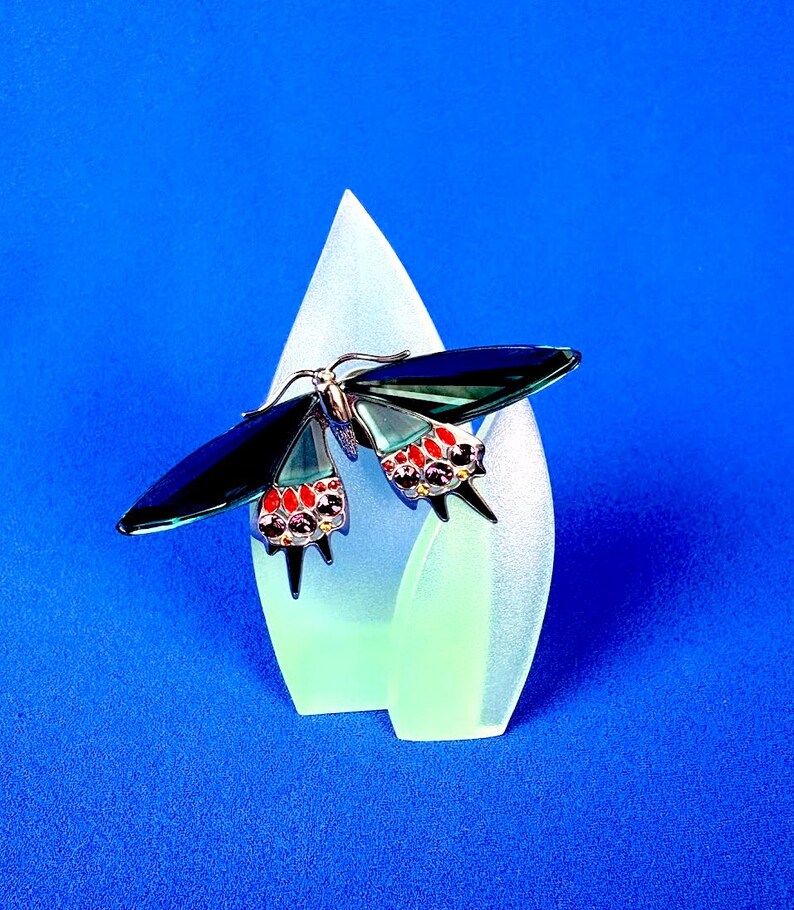 Puede incluir: Broche decorativo de mariposa con alas azul oscuro y detalles rojos y morados. El broche est&aacute; colocado sobre una pantalla en forma de l&aacute;grima verde claro sobre un fondo azul.