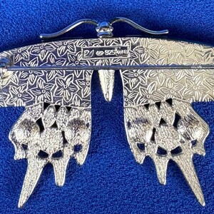 Puede incluir: Broche de mariposa plateada con detalles intrincados. El broche presenta un patr&oacute;n floral en las alas y un cierre de alfiler. Las alas de la mariposa est&aacute;n abiertas, sobre un fondo azul.