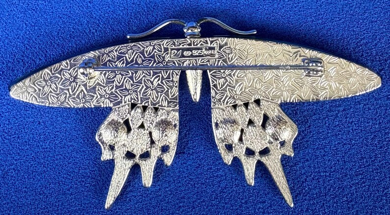 Puede incluir: Broche de mariposa plateada con detalles intrincados. El broche presenta un patr&oacute;n floral en las alas y un cierre de alfiler. Las alas de la mariposa est&aacute;n abiertas, sobre un fondo azul.