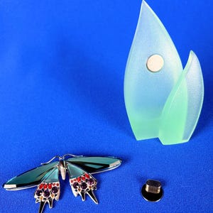 Puede incluir: Un broche de mariposa plateado con alas verde azulado y detalles rojos y morados. Un objeto decorativo azul claro y verde con un im&aacute;n y un peque&ntilde;o objeto negro y plateado sobre un fondo azul.