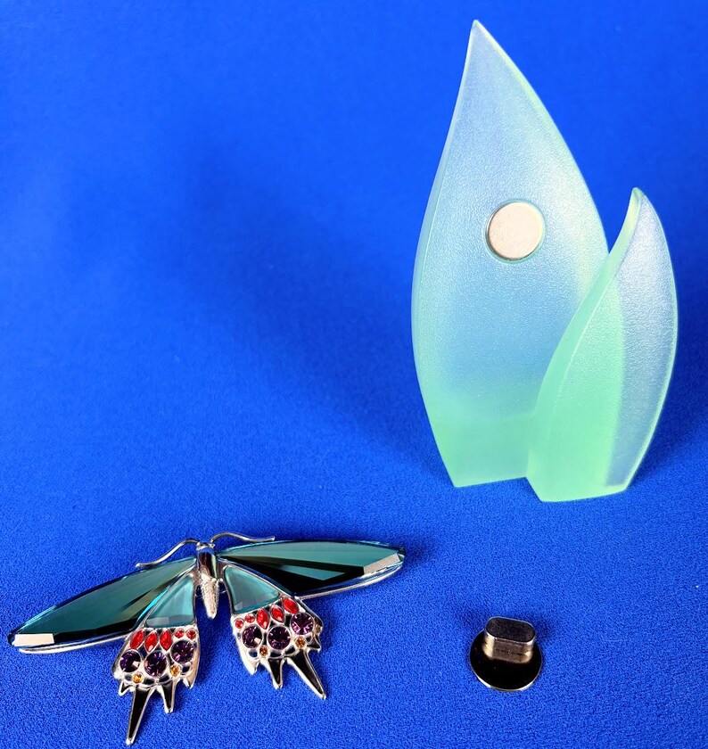 Puede incluir: Un broche de mariposa plateado con alas verde azulado y detalles rojos y morados. Un objeto decorativo azul claro y verde con un im&aacute;n y un peque&ntilde;o objeto negro y plateado sobre un fondo azul.