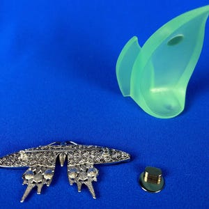 Puede incluir: Un broche de mariposa plateado con intrincados detalles florales, un elemento decorativo verde transl&uacute;cido y un cierre de alfiler dorado. Los art&iacute;culos est&aacute;n dispuestos sobre un fondo azul.