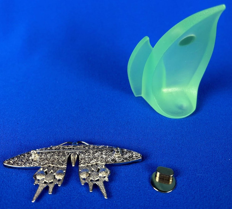 Puede incluir: Un broche de mariposa plateado con intrincados detalles florales, un elemento decorativo verde transl&uacute;cido y un cierre de alfiler dorado. Los art&iacute;culos est&aacute;n dispuestos sobre un fondo azul.