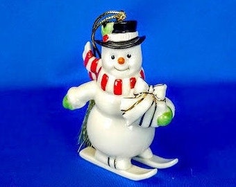 May include: A white porcelain snowman ornament wearing a red and white striped scarf and a black top hat. The snowman is skiing down a white slope.