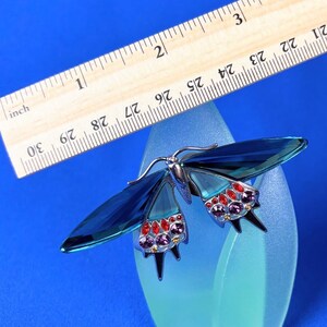 Puede incluir: Broche de mariposa azul con cuerpo de metal plateado y alas. Las alas son de cristal azul y est&aacute;n adornadas con cristales rojos y morados. Una regla de madera est&aacute; colocada encima.