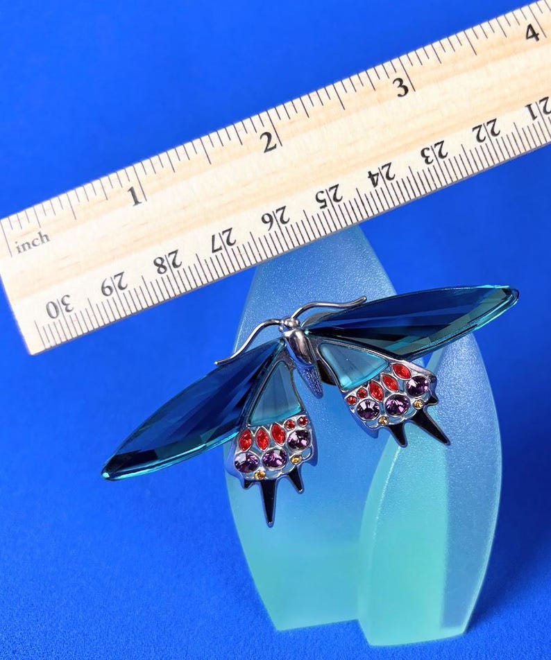 Puede incluir: Broche de mariposa azul con cuerpo de metal plateado y alas. Las alas son de cristal azul y est&aacute;n adornadas con cristales rojos y morados. Una regla de madera est&aacute; colocada encima.