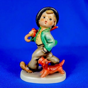 May include: A porcelain figurine of a boy wearing a green jacket and black hat, holding an umbrella and walking with a red dog.