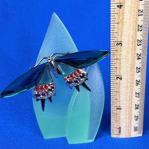 Puede incluir: Una figura decorativa de mariposa con alas azules y detalles de joyas, montada sobre una base de vidrio esmerilado. La envergadura de la mariposa es de aproximadamente 5 cm. La base es de color azul claro y el fondo es azul liso.