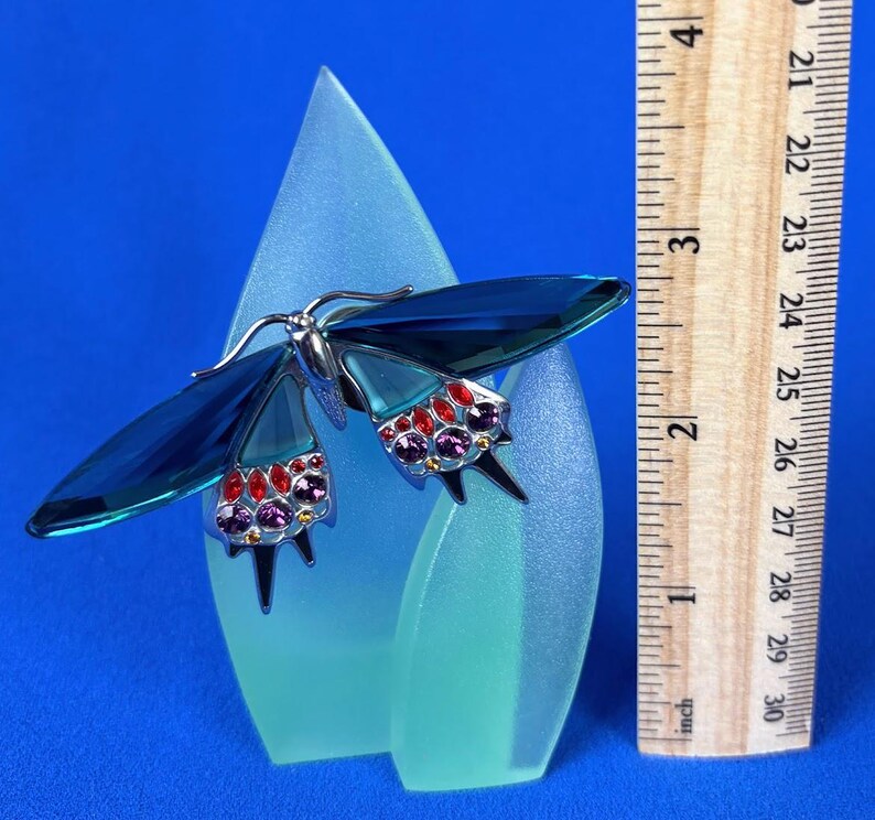 Puede incluir: Una figura decorativa de mariposa con alas azules y detalles de joyas, montada sobre una base de vidrio esmerilado. La envergadura de la mariposa es de aproximadamente 5 cm. La base es de color azul claro y el fondo es azul liso.