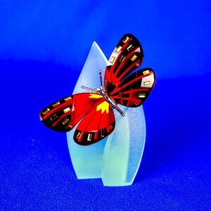 May include: A red and black butterfly figurine with a yellow center, perched on a frosted glass stand. The stand is shaped like two overlapping teardrops.